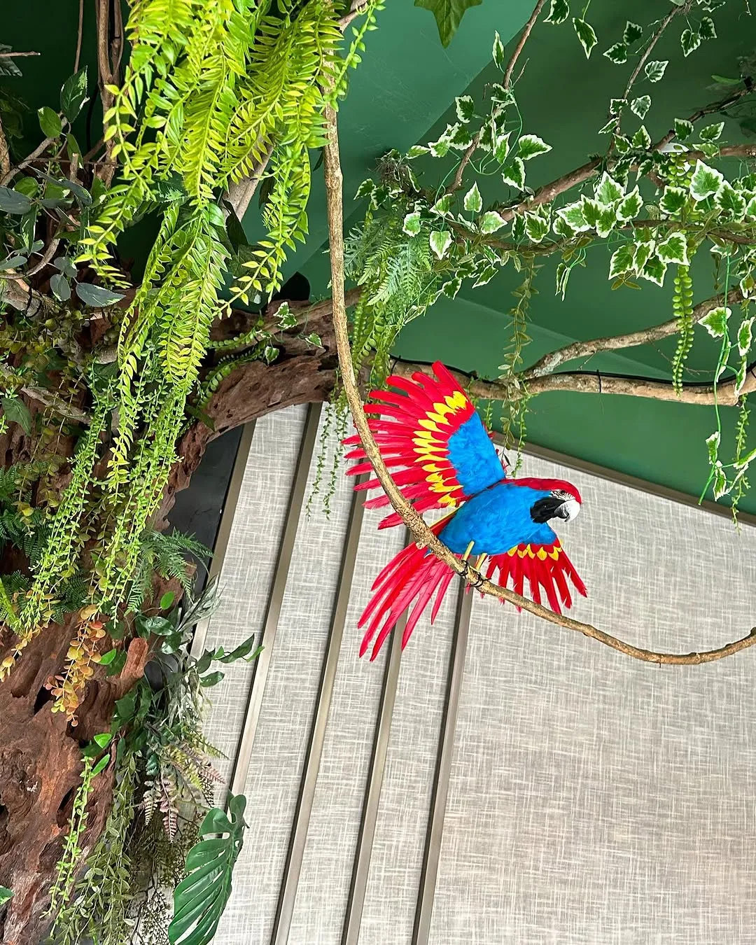 Escultura de arara colorida sobre parede de vegetação, junto à escada de acesso