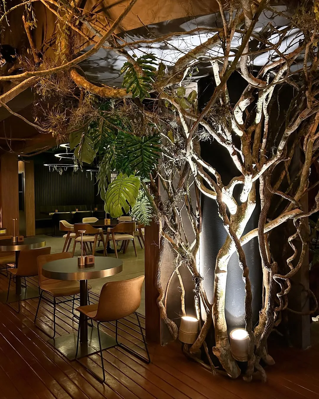 Salão do Mango com galhos de árvore formando um biombo estrutural, iluminados de baixo para cima por balizadores no piso de madeira
